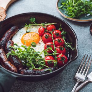 Breakfast time, fried egg with sausages and cherry tomatoes in a black iron pan, served microgreens.